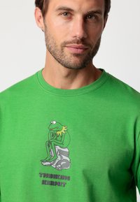 Camiseta de algodón verde con un estampado de la pantalla que muestra a Kermit la Rana sentado en una roca, con el texto "KERMIT PENSATIVO" abajo en letras negras y en negrita.