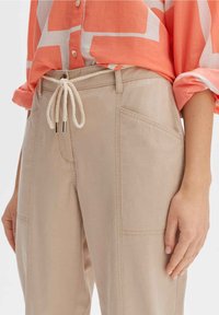 Beige Hosen mit einem Kordelzugbund mit metallenen Spitzen, seitlichen Taschen und einem entspannten Schnitt, kombiniert mit einem orangefarbenen gemusterten Oberteil.