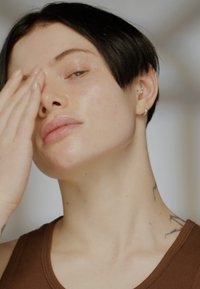 Zbliżenie na osobę z krótkimi czarnymi włosami, ubraną w brązowy top na ramiączkach. Ma gładką skórę, lekkie makijaż i widoczne kolczyki w uszach.