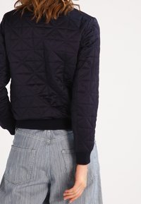 Chaqueta acolchada azul marino con un patrón geométrico, puños acanalados y un diseño cropped, llevada sobre pantalones de pierna ancha en azul claro.