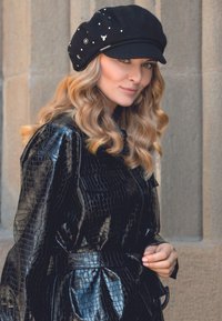 Cappotto nero, lucido, con texture a stampa coccodrillo, cinturato in vita e con ampie rever; berretto di lana nera decorato con accenti decorativi e perline.
