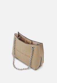 Beige konstläderhandväska med silverfärgad kedjeaxelrem, präglad "Calvin Klein Jeans"-logotyp, strukturerad kropp och öppet topplock.