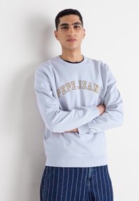 Sudadera azul claro con cuello redondo y "PEPE JEANS" impreso en beige. Presenta puños y dobladillo acanalados, combinada con pantalones navy a rayas.