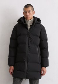 Schwarze Puffermantel mit Kapuze, mit Reißverschluss, gesteppter Optik und zwei Seitentaschen. Getragen über einem leichten Pullover.