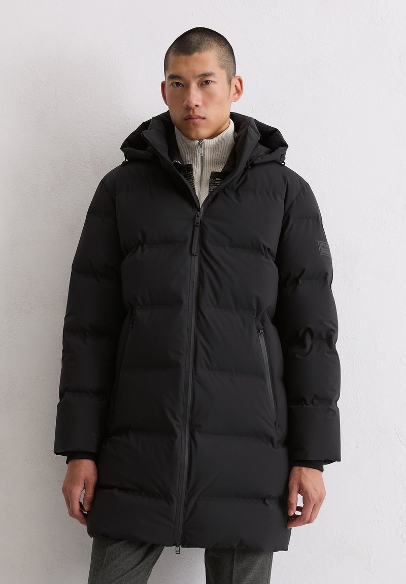 Schwarze Puffermantel mit Kapuze, mit Reißverschluss, gesteppter Optik und zwei Seitentaschen. Getragen über einem leichten Pullover.