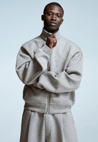 Giovane uomo in felpa grigia chiara oversize con zip e pantaloni abbinati, che tiene la cerniera vicino al colletto, in piedi contro uno sfondo chiaro.