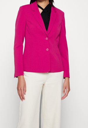 Vrouw draagt een felroze blazer met twee knopen over een zwarte top en witte wijde broekspijpen, staand tegen een effen achtergrond.