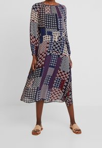Femme portant une robe à manches longues, longueur genoux, avec un motif patchwork multicolore et des sandales plates beige à bouts ouverts.