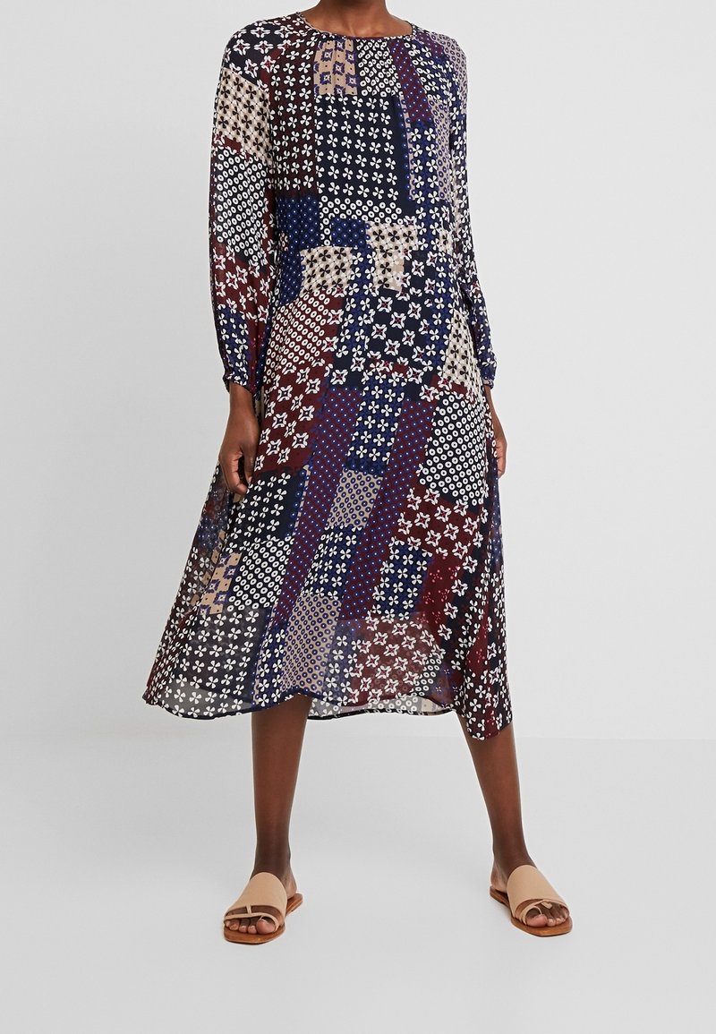 Femme portant une robe à manches longues, longueur genoux, avec un motif patchwork multicolore et des sandales plates beige à bouts ouverts.