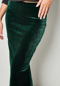 Gonna maxi in velluto verde con silhouette aderente, morbida al tatto e dettagli drappeggiati, che mettono in evidenza il suo fluire continuo dall'anca all'orlo.