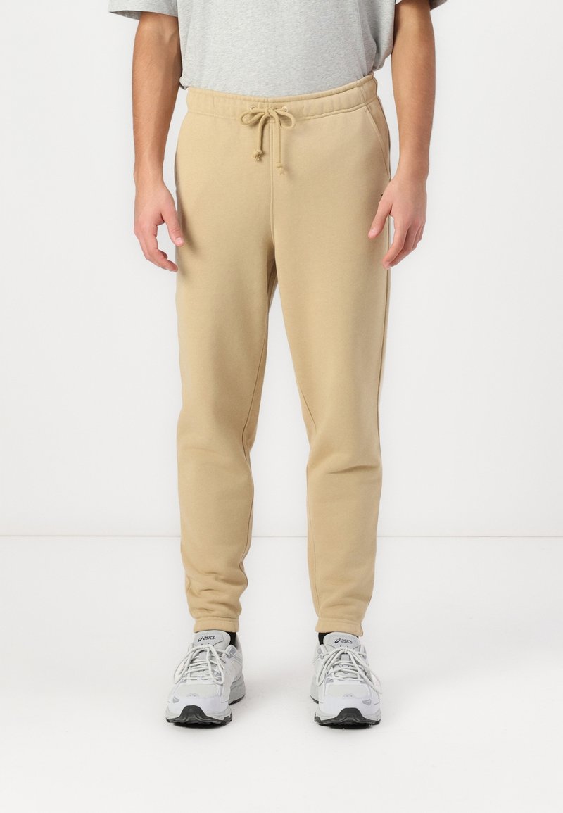 Béžové slim-fit tepláky s elastickým páskem, vyrobené z jemné tkaniny. Disponují bočními kapsami a zúženými nohavicemi, kombinované se šedými teniskami.