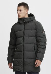 Tamnozelena puffer jakna s kapuljačom, s patent zatvaračem, bočnim džepovima i kviltiranim uzorkom. Glatka tekstura s suptilnim sjajem.