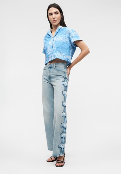 Γυναίκα που φοράει μπλε κοντομάνικο πουκάμισο με λουλουδάτο μοτίβο και ανοιχτό μπλε τζιν με μπλε λουλουδάτο σχέδιο στο πλάι, στέκεται με το ένα χέρι στη μέση.