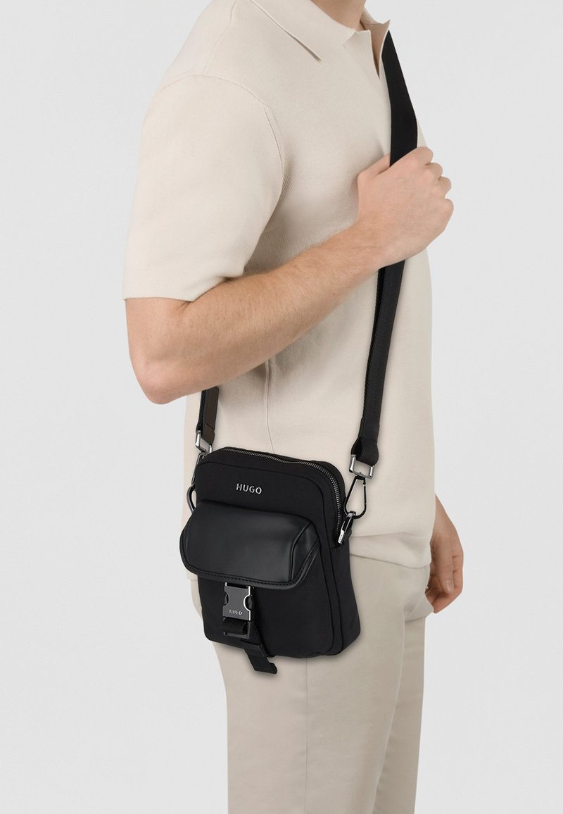 Schwarze Crossbody-Tasche aus strukturiertem Stoff mit glatten Lederakzenten, ausgestattet mit einem Vorderklappendeckel, Reißverschluss und verstellbarem Tragegurt.