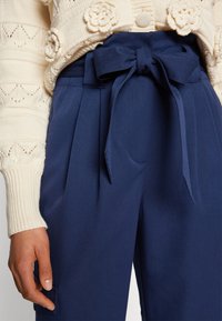 Navy blue trousers with pleats, a tied waist detail, and a smooth texture. Paired with a cream, patterned knitted sweater.