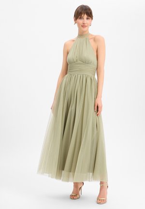 Femme portant une robe sans manches vert olive avec un col halter, un corsage plissé et une jupe fluide, associée à des talons hauts dorés à brides.