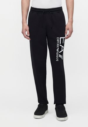 Mand iført sorte Emporio Armani joggingbukser med hvidt logo på venstre lår og sorte sneakers med hvide såler.