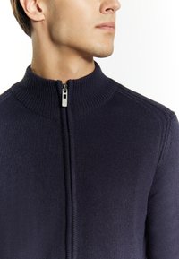 Jūras spēku zila adīta jaka ar augstu apkakli un priekšējo rāvējslēdzēju. Struktūrēta auduma virsma ar līnijām apkakles un plecu daļā.