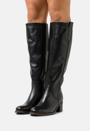 Bottes en cuir noires montantes jusqu'aux genoux avec talons carrés portées sur des jambes nues, avec fermetures éclair latérales et détails de coutures.
