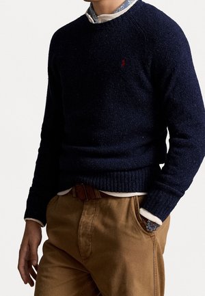 Man draagt een donkerblauwe trui over een licht patroonhemd en beige broek, met één hand in de zak en een horloge zichtbaar.