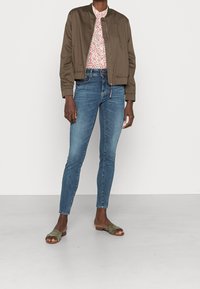 Chaqueta bomber verde oliva, camisa floral, jeans ajustados azules y sandalias oliva. Tela suave, ajuste casual, con detalles de cremallera y suela plana.