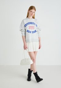 Jovem mulher vestindo um sweatshirt cinzento "Universidade de NYC Nova Iorque", saia branca, botas pretas, segurando uma bolsa branca, de pé contra um fundo simples.