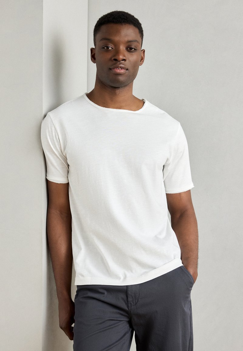 T-shirt bianca in cotone con scollatura rotonda e maniche corte, caratterizzata da una texture sottile. Indossata con pantaloni corti grigio scuro.