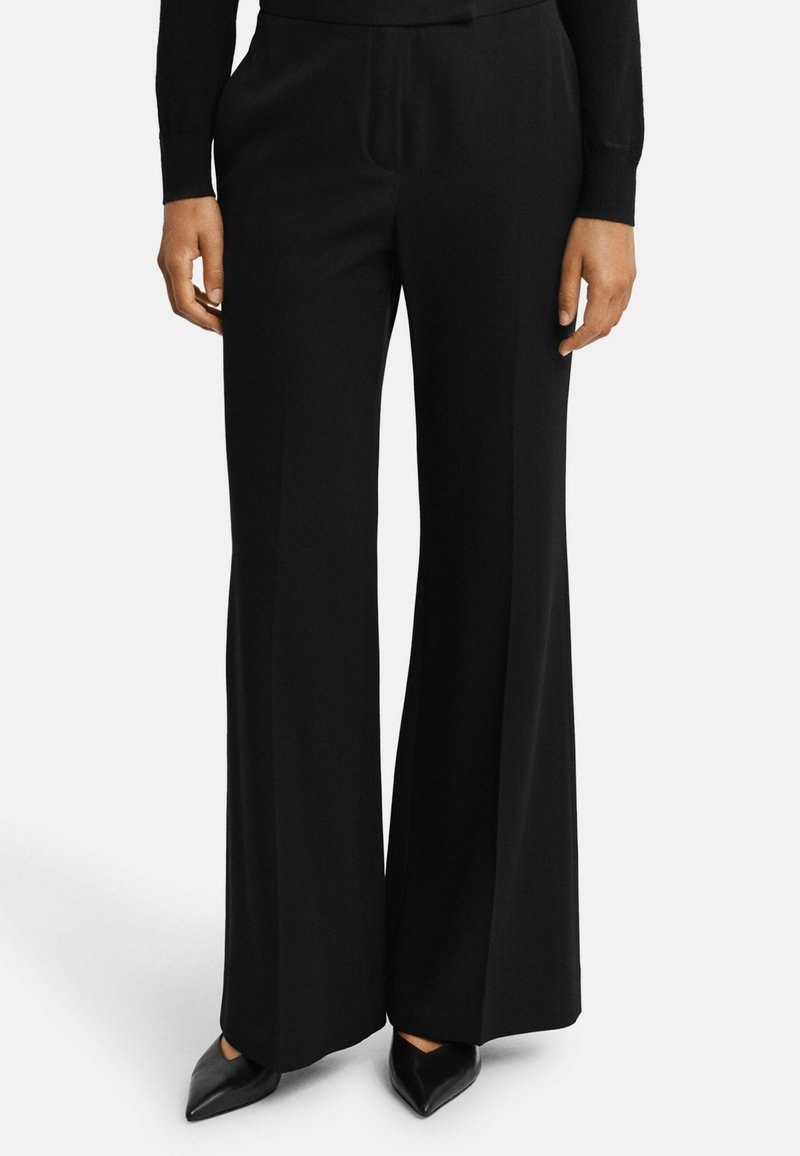 Zwarte wijde broek met een gladde textuur, met een getailleerde pasvorm en plooien aan de voorkant, gecombineerd met spitse zwarte hakken.