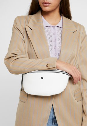 Frau trägt ein beiges Nadelstreifen-Blazer und ein lavendelfarbenes Spitzen-Poloshirt, hält eine weiße, mit Reißverschluss versehene Bauchtasche quer über den Oberkörper.