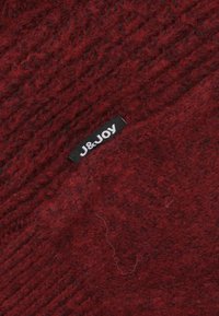 Getextureerde rode gebreide stof met een label met "J&JOY" in het wit, tegen een donkerder rode achtergrond, die een rijke, warme kleur toont.