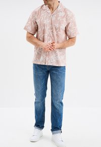 Camisa de manga corta, de color rosa claro con patrones florales blancos, combinada con jeans de mezclilla azules y zapatillas blancas. Diseño casual y de corte relajado.