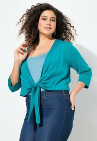 Cardigan cropped teal avec un nœud à l'avant, manches longues, associé à un haut bleu clair et un jean bleu foncé, sur fond clair.