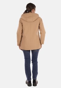 Giacca impermeabile beige con cappuccio, texture liscia e taglio dritto, abbinata a jeans scuri e scarpe nere con lacci.