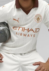 Valkoinen jalkapallopaita ruskealla kauluksella, jossa on teksturoitu muotoilu, joukkueen logo ja oranssi sponsori-brändäys. Pelaaja pitää harmaata jalkapalloa.