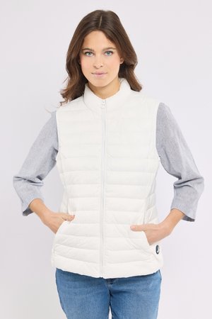 Kvinde iført en hvid quiltet ærmeløs vest over en grå langærmet skjorte og blå jeans med hænderne i vestens lommer.