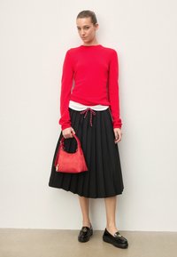 Femme portant un haut rouge à manches longues, une jupe midi plissée noire et des mocassins noirs, tenant un sac à main rouge contre un mur uni.