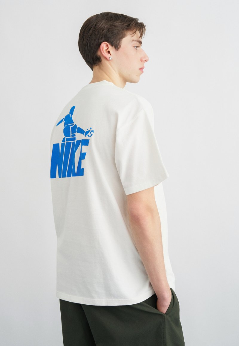 Junger Mann mit kurzem braunem Haar trägt ein weißes Nike-T-Shirt mit großem blauem Logo und Grafik auf der Rückseite, kombiniert mit dunkelgrünen Hosen.
