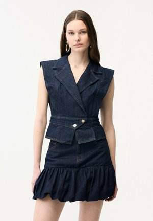 Kobieta ubrana w ciemny, bezrękawnik z denimu z złotymi guzikami oraz pasującą, krótką, falbanianą spódnicę, dużą złotą kolczyki koła, neutralne tło.