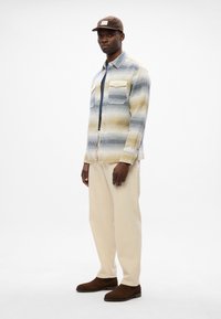 Herrenhemdjacke mit einem Karomuster in Blau, Gelb und Weiß. Verfügt über zwei Brusttaschen, Knöpfe und lange Ärmel; kombiniert mit cremefarbenen Hosen und braunen Schuhen.