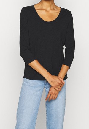 Personne portant un t-shirt noir à col rond avec des manches trois-quarts et un jean bleu clair, debout les mains jointes devant.