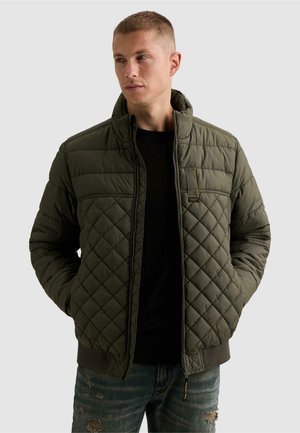 Homme portant une veste matelassée vert olive sur une chemise noire et un jean déchiré, mains dans les poches, regardant vers sa gauche.