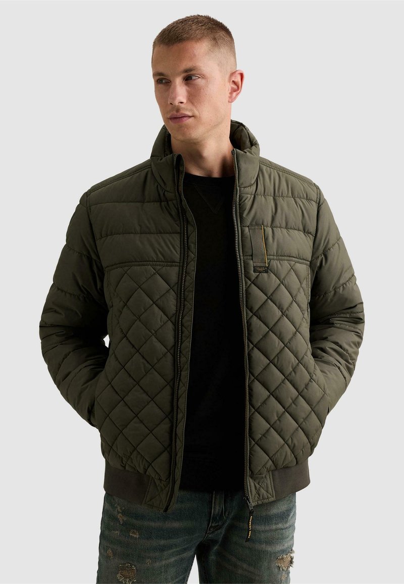 Homme portant une veste matelassée vert olive sur une chemise noire et un jean déchiré, mains dans les poches, regardant vers sa gauche.