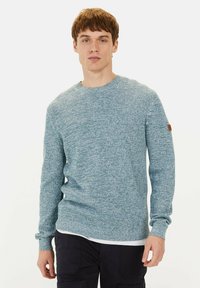 Textured blue knit sweater with long sleeves, round neckline, and a small brown logo patch on the left arm, worn over a white shirt.