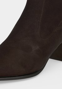 Gros plan d’une botte cheville en daim marron foncé, bout arrondi, coutures visibles et petit talon carré sur fond clair.