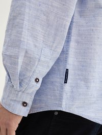 Light blue button-up shirt with a textured fabric, featuring small repeating patterns and dark brown buttons on the cuff. Brand tag visible.