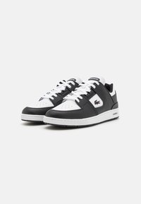 Zapatillas en blanco y negro con una parte superior de cuero, puntera perforada, cordones blancos y un panel lateral de textura negra con la marca Lacoste.