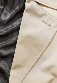 Beige stof met een gladde textuur naast zwart, gemustreerd materiaal. Heeft stiksels en een decoratieve lusdetail aan de rand.