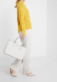 Sac fourre-tout blanc avec des accents noirs et des poignées hexagonales, associé à un pull en tricot jaune et un pantalon large beige clair. Talons hauts roses.