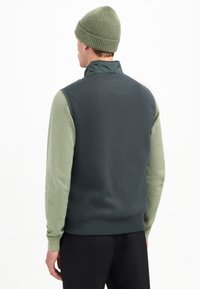 Donkergroene mouwloze vest met textuur in lichtgroene mouwen. Heeft een hoge kraag en geribbelde zoom. Gecombineerd met een bijpassende gebreide beanie.