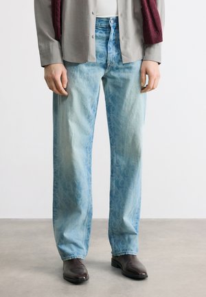 Personne portant un jean bleu clair à motifs, coupe droite, des chaussures marron foncé, une chemise grise et un pull bordeaux posé sur les épaules.